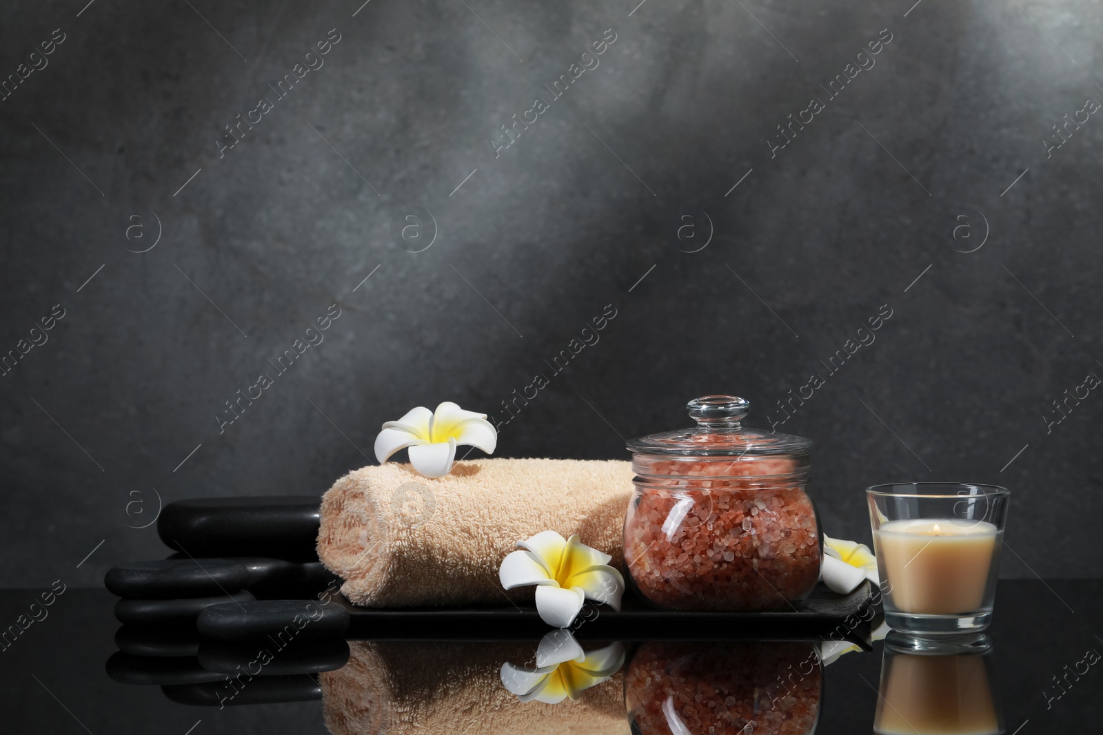 Photo of Glass jar with natural sea salt, beautiful flowers, candle and spa stones on mirror surface