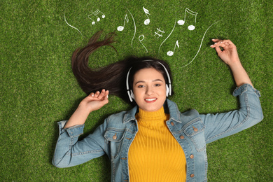 Young woman listening to music through headphones on green grass, top view