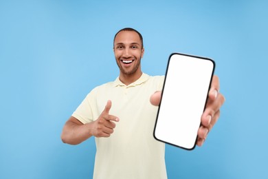 Young man showing smartphone in hand and pointing at it on light blue background