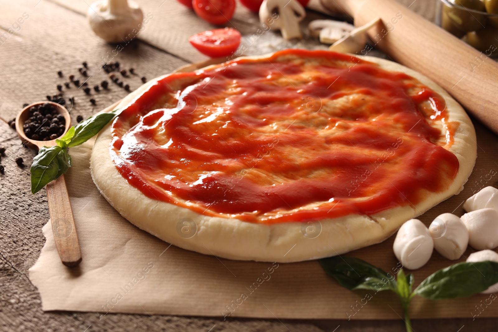 Photo of Pizza dough smeared with tomato sauce and products on wooden table, closeup