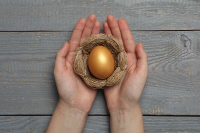 Photo of Woman holding nest with shiny golden egg at grey wooden table, top view