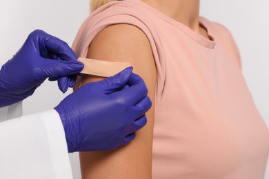 Nurse sticking adhesive bandage on woman's arm after vaccination on light background, closeup