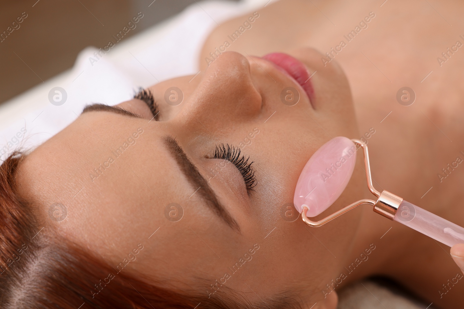 Photo of Young woman receiving facial massage with rose quartz roller in beauty salon, closeup