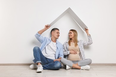 Young family housing concept. Pregnant woman with her husband sitting under cardboard roof on floor indoors