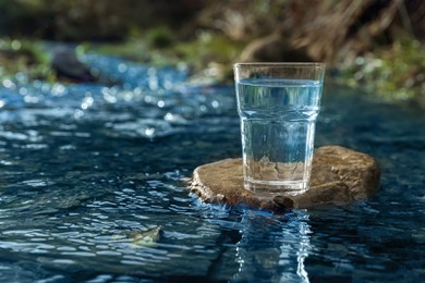 Photo of Glass of fresh water on stone near river outdoors. Space for text