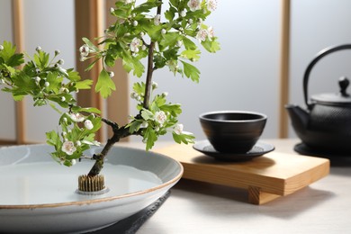 Photo of Stylish ikebana as house decor. Beautiful fresh branch with flowers and tea set on white table, space for text