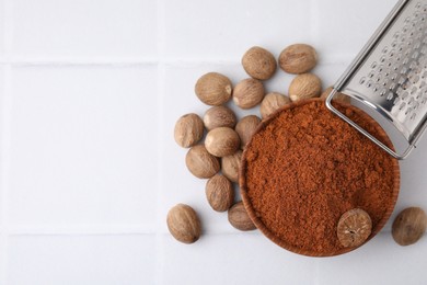 Photo of Nutmeg powder in bowl, seeds and grater on white tiled table, flat lay. Space for text