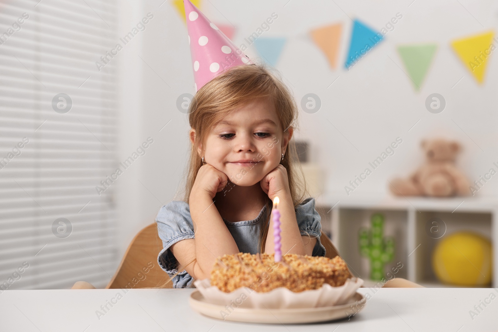 Photo of Cute girl in party hat with birthday cake at table indoors