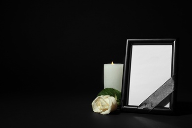 Photo of Funeral photo frame with ribbon, white rose and candle on dark table against black background. Space for design