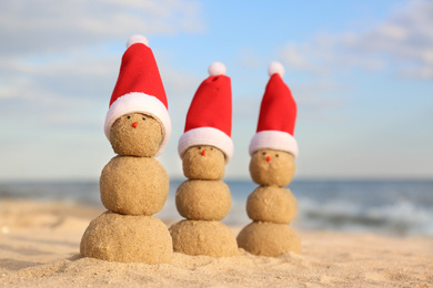 Photo of Snowmen made of sand with Santa hats on beach near sea. Christmas vacation