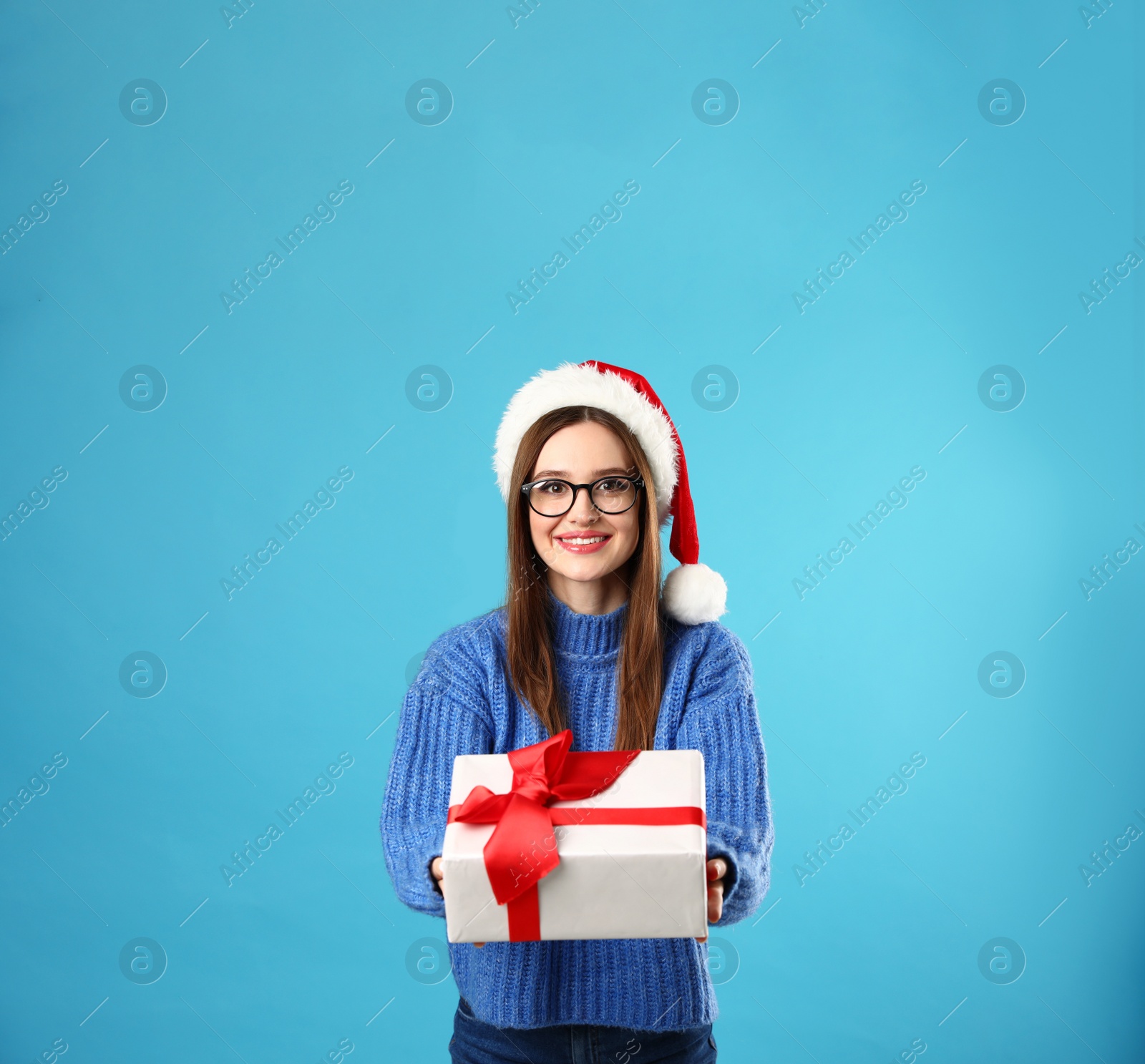 Photo of Young woman in Christmas sweater with Santa hat and gift box on blue background