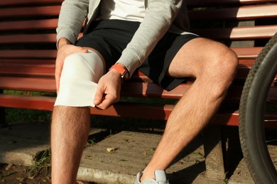 Photo of Man applying bandage onto his knee on wooden bench outdoors, closeup