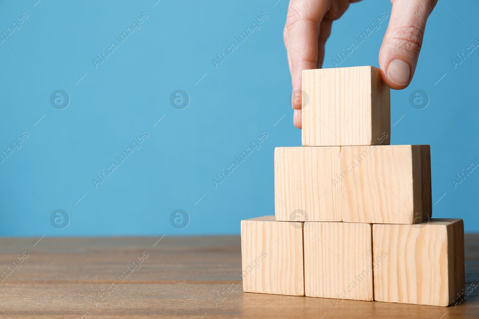 Photo of Woman building pyramid of cubes on wooden table against light blue background, closeup. Space for text