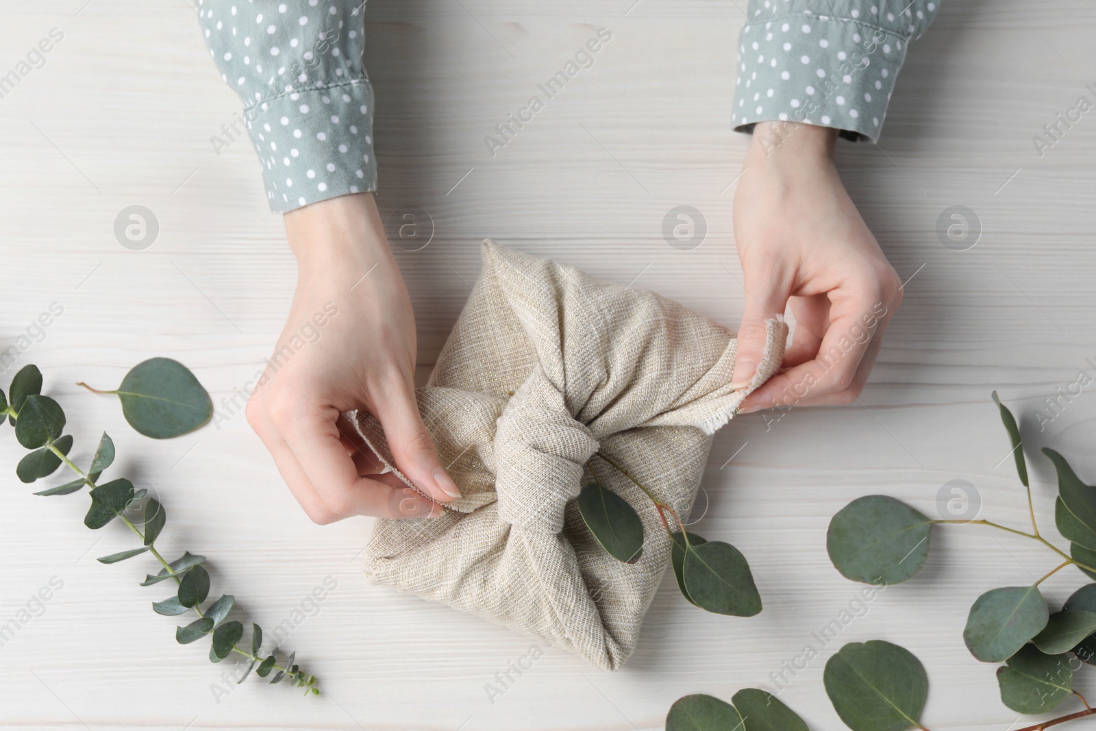 Photo of Furoshiki technique. Woman wrapping gift in fabric at white wooden table, top view