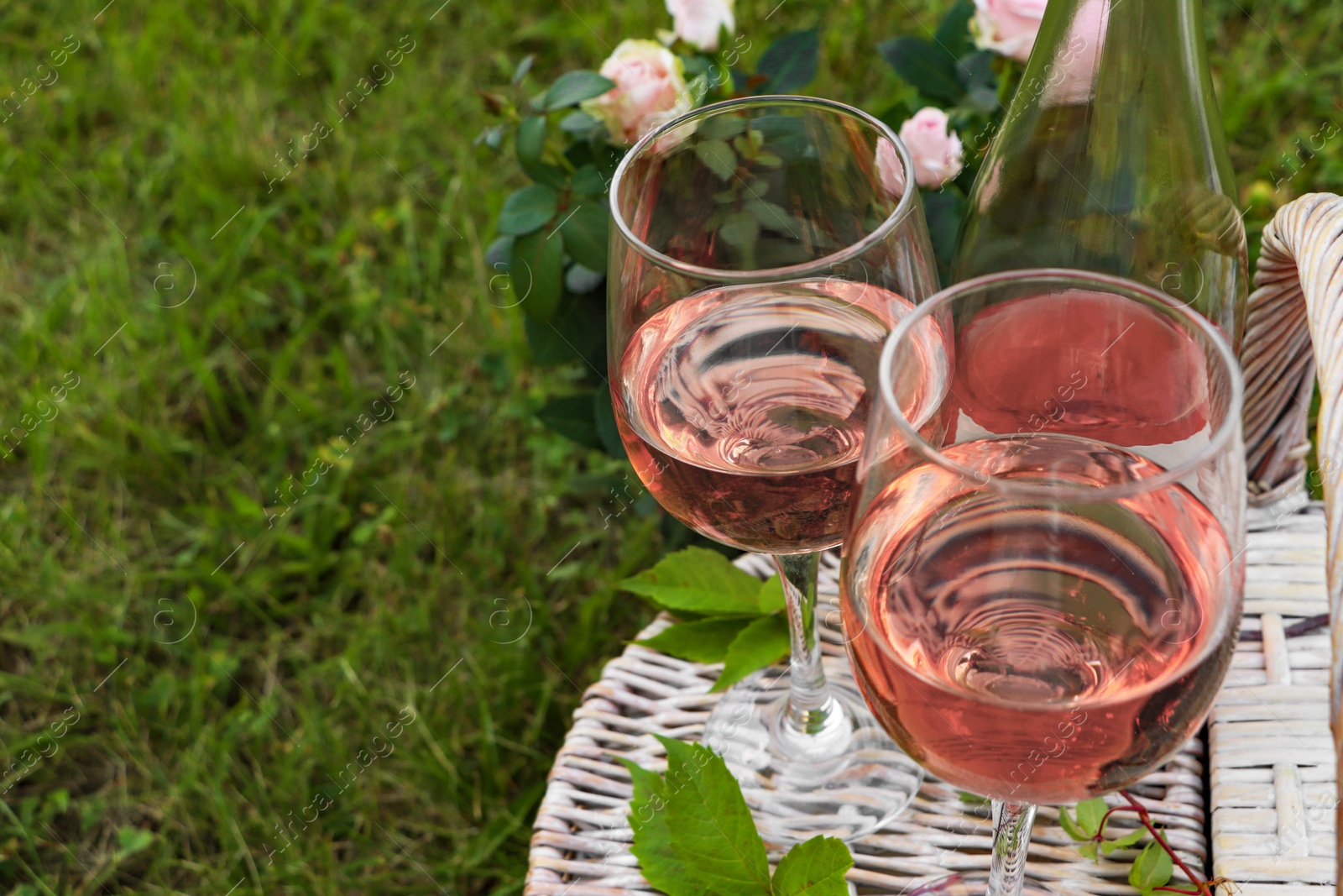 Photo of Glasses and bottle of delicious rose wine on picnic basket outdoors, closeup. Space for text