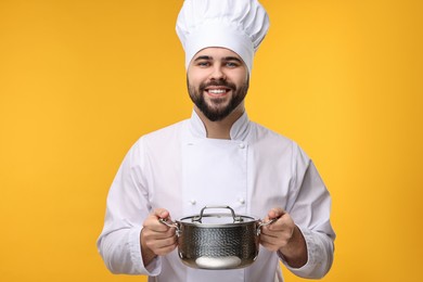 Photo of Happy young chef in uniform holding cooking pot on orange background