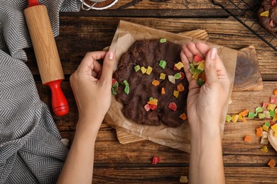Photo of Woman adding candied fruits to unbaked chocolate cookie at wooden table, top view