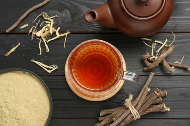 Aromatic licorice tea in cup, teapot, dried sticks of licorice root and powder on black wooden table, flat lay