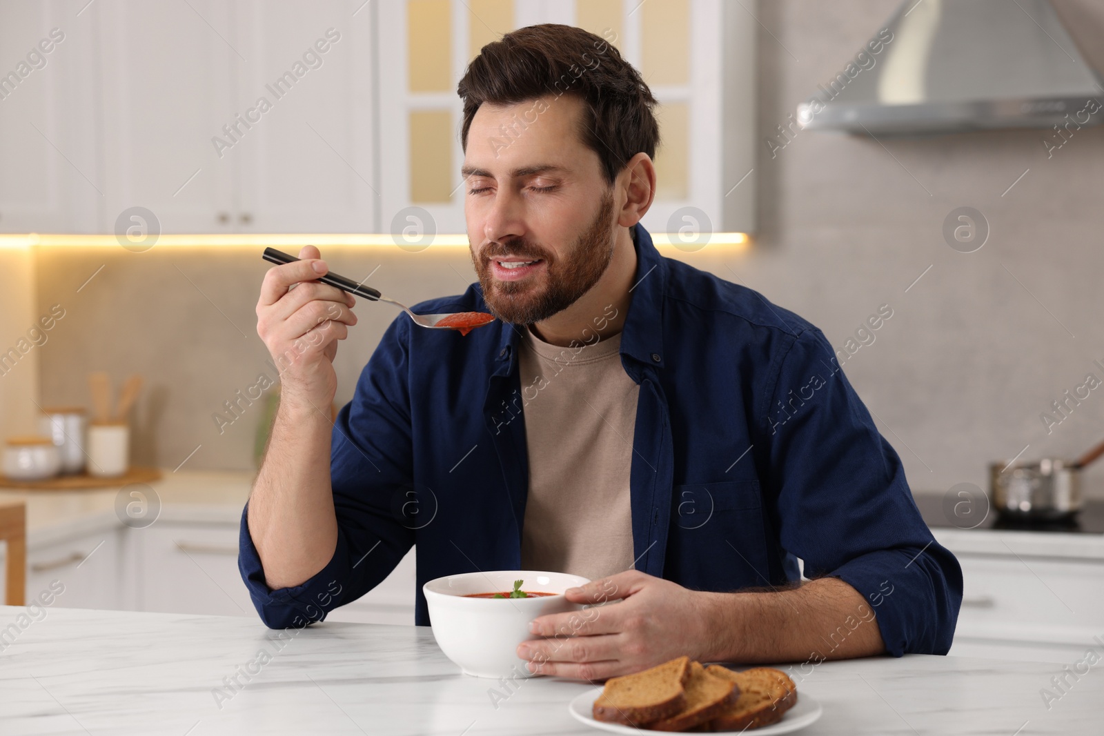 Photo of Man eating delicious tomato soup at light marble table in kitchen