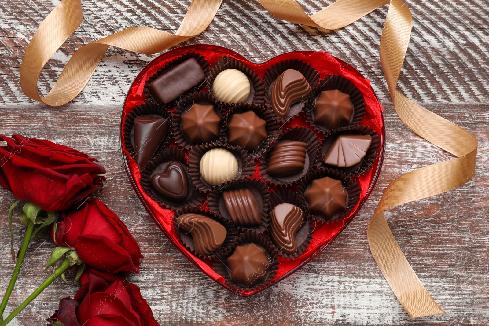 Photo of Heart shaped box with delicious chocolate candies, roses and ribbon on wooden table, flat lay