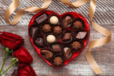 Photo of Heart shaped box with delicious chocolate candies, roses and ribbon on wooden table, flat lay