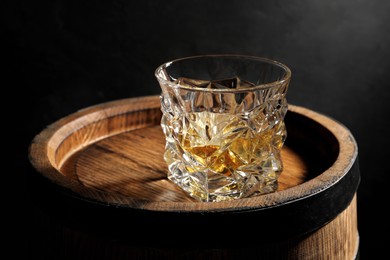 Photo of Glass of whiskey on wooden barrel against black background, closeup