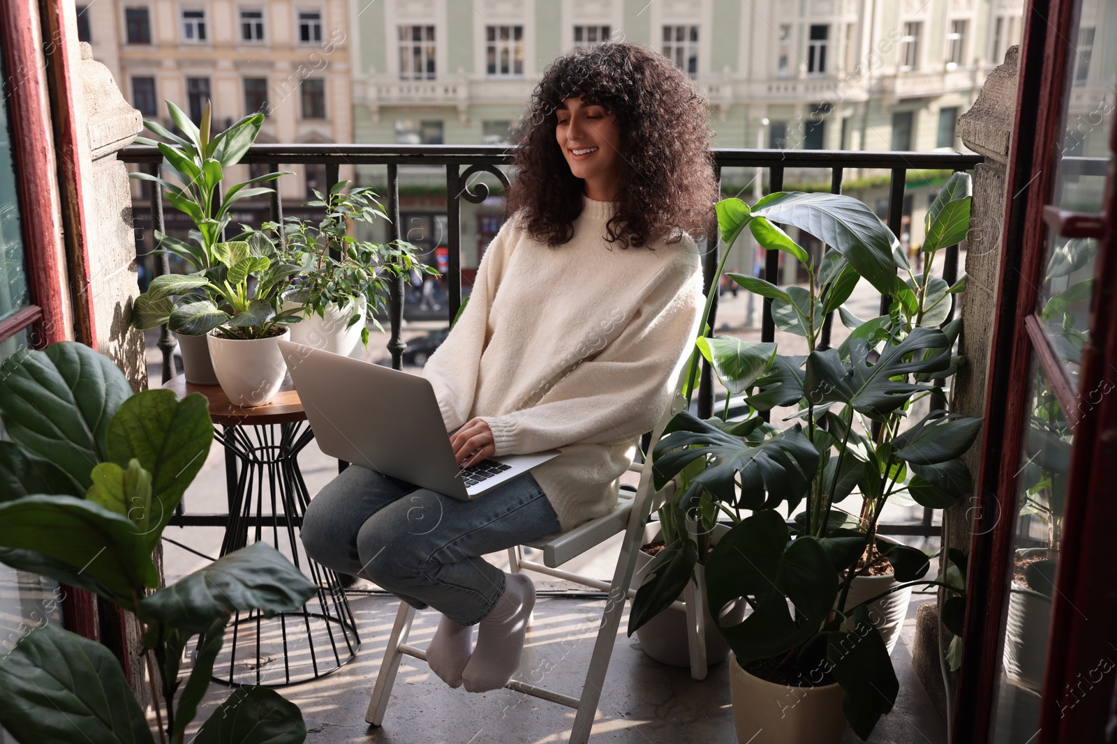 Photo of Beautiful young woman using laptop surrounded by green houseplants on balcony