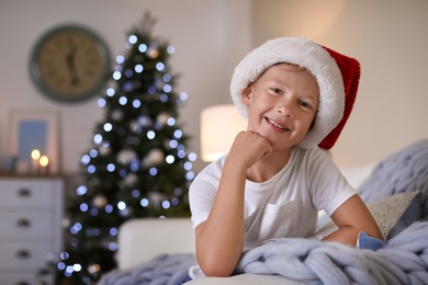 Cute little child in Santa hat sitting on sofa at home. Christmas celebration