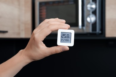 Photo of Woman holding digital hygrometer with thermometer in kitchen, closeup