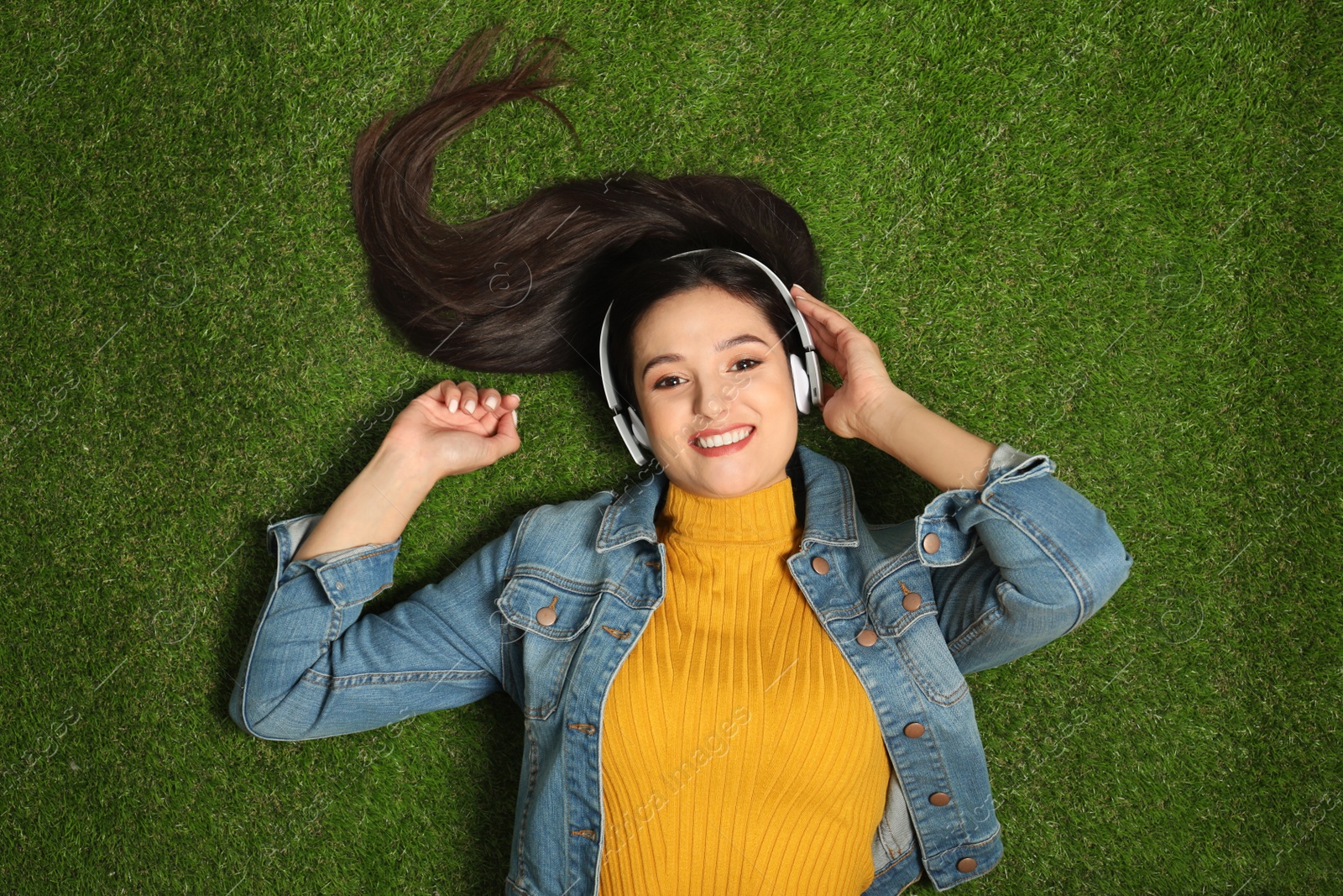 Photo of Young woman listening to audiobook while lying on grass, top view