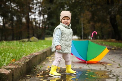 Photo of Cute little girl having fun in puddle near colorful umbrella outdoors