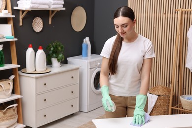 Photo of Beautiful woman with spray bottle and microfiber cloth cleaning white table in laundry room