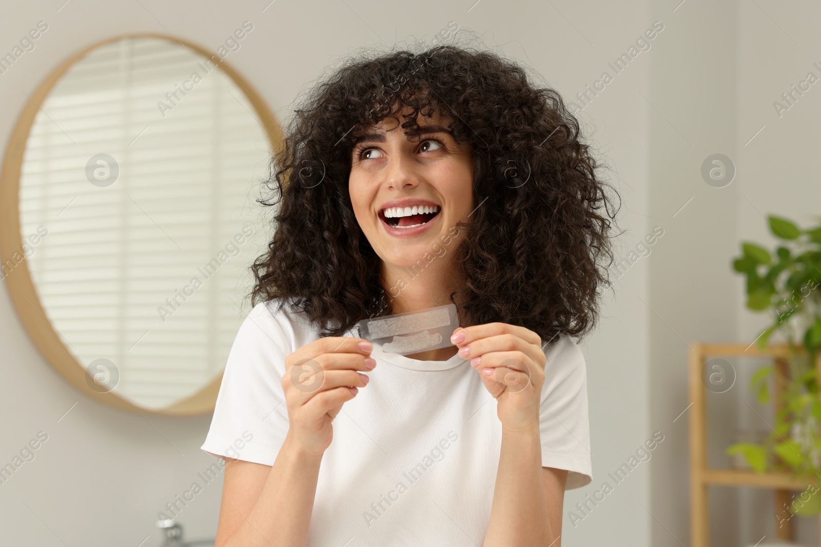 Photo of Young woman holding teeth whitening strips in bathroom