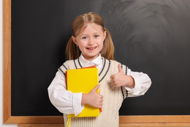 Happy schoolgirl with backpack and books showing thumb up gesture near blackboard