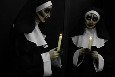 Photo of Scary devilish nun with burning candle near mirror on black background. Halloween party look
