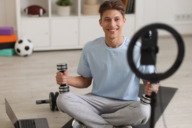 Photo of Smiling sports blogger holding dumbbells while streaming online fitness lesson at home