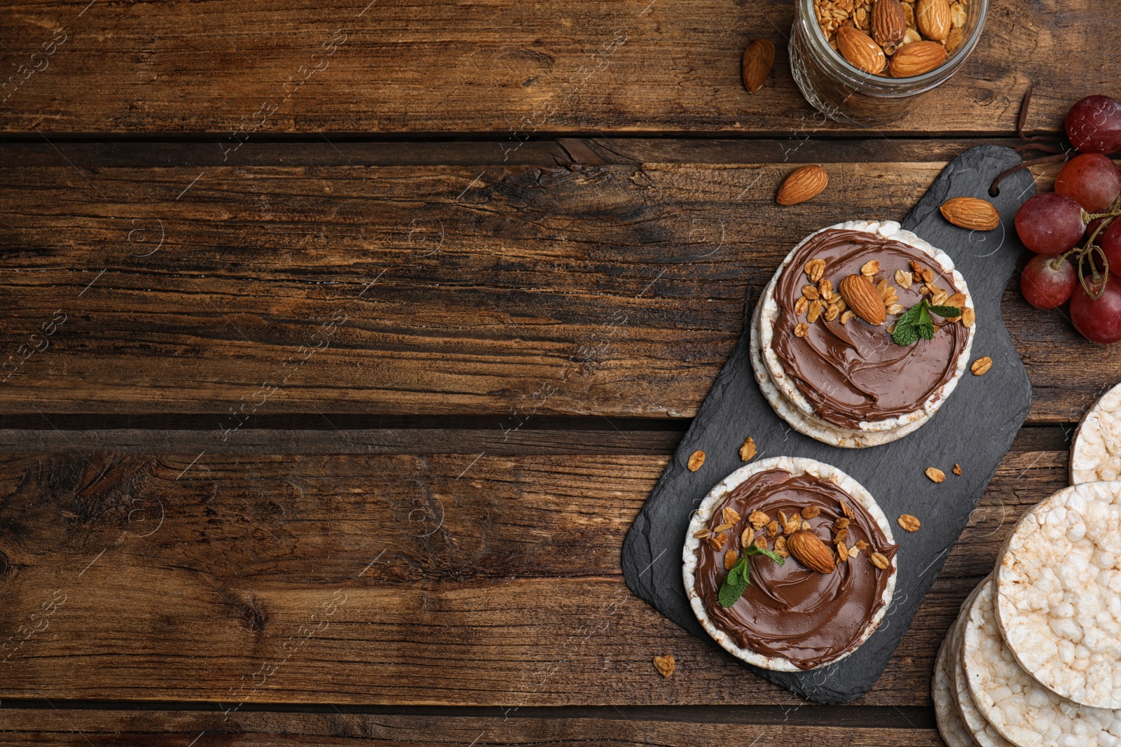 Photo of Puffed rice cakes with chocolate spread, nuts and mint on wooden table, flat lay. Space for text