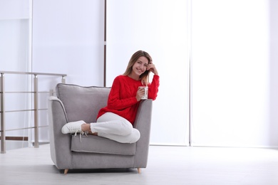 Photo of Attractive young woman with cup of tea in armchair indoors. Space for text