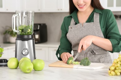 Photo of Young woman cutting broccoli for smoothie at white table in kitchen, closeup