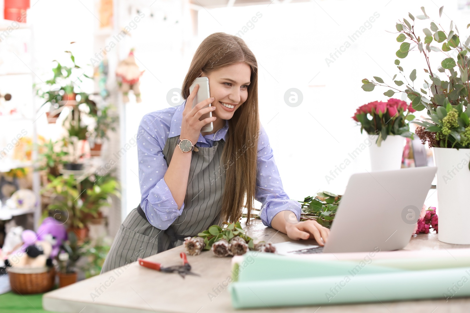 Photo of Young florist working with laptop and talking on mobile phone in flower shop