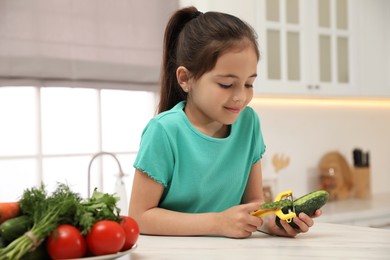 Photo of Little girl peeling cucumber at table in kitchen. Preparing vegetable