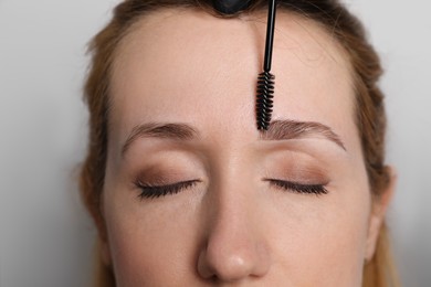 Photo of Beautician brushing woman's eyebrows before tinting on light grey background, closeup
