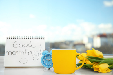 Delicious coffee, alarm clock, beautiful flowers and GOOD MORNING wish on light windowsill