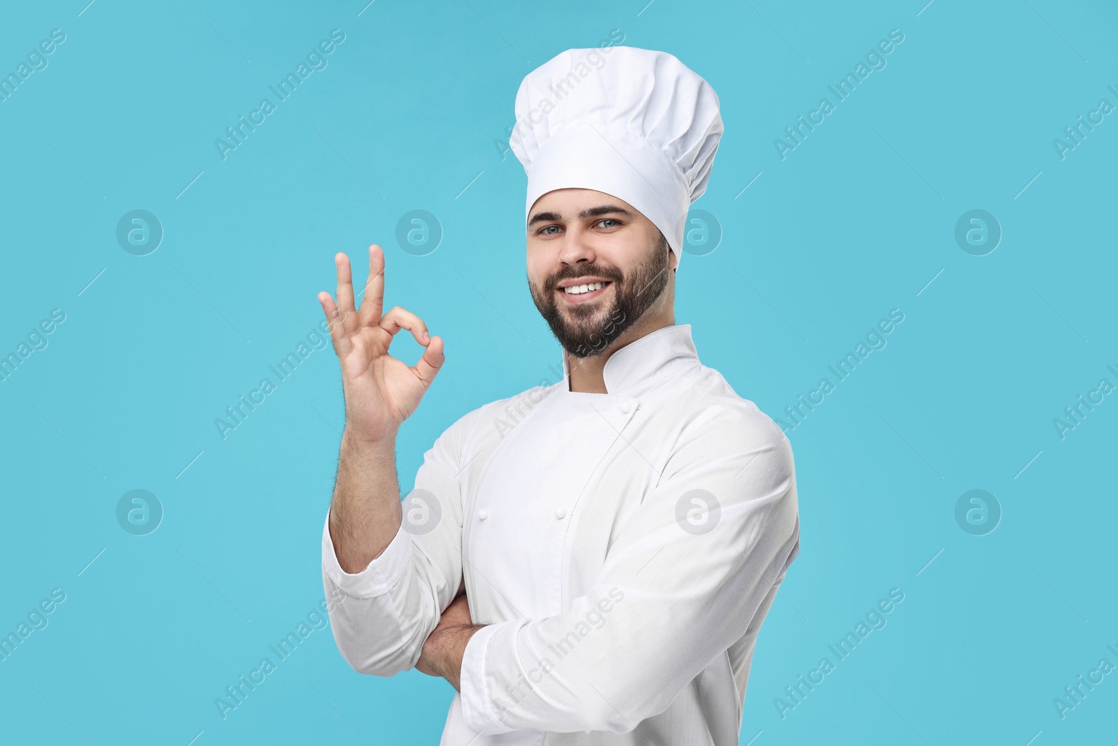 Photo of Happy young chef in uniform showing ok gesture on light blue background