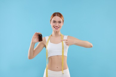 Photo of Happy young woman with bottle of pills and measuring tape on light blue background. Weight loss