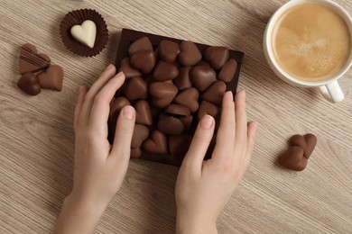 Woman with box of heart shaped chocolate candies and coffee at wooden table, top view