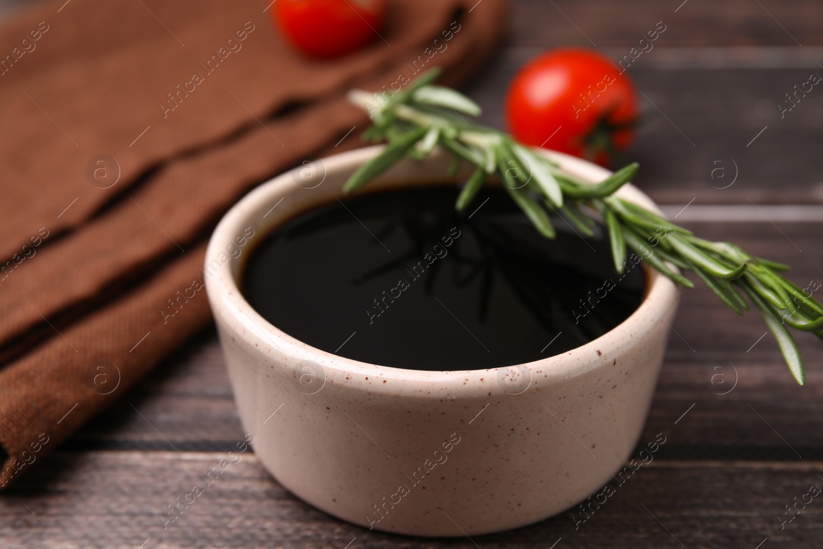 Photo of Bowl with balsamic vinegar, rosemary and tomatoes on wooden table, closeup