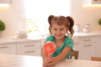 Photo of Little girl enjoying air flow from portable fan at table in kitchen. Summer heat