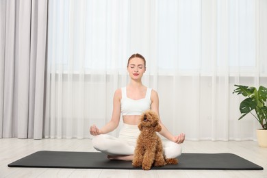 Photo of Young woman practicing yoga on mat with her cute dog indoors