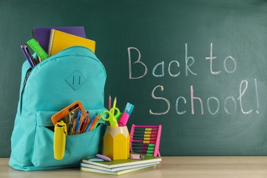 Photo of Bright backpack with school stationery on wooden table near green chalkboard. Back to School